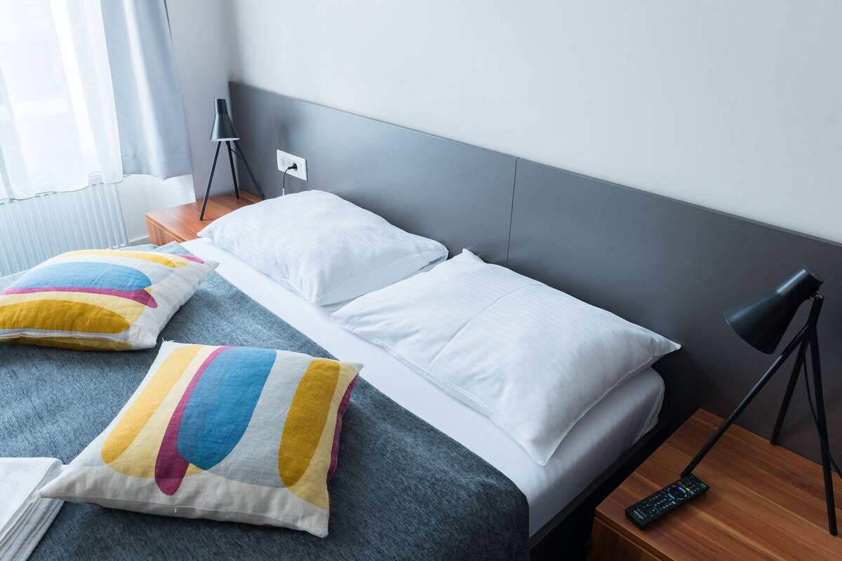 A neatly made bed with white and colorful pillows, a remote control on a wooden nightstand, and a lamp beside the bed.