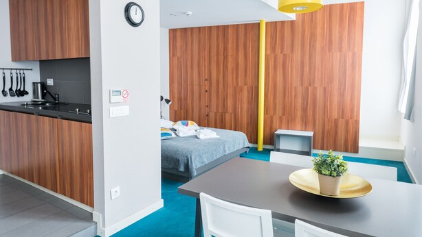 A modern hotel room with a kitchenette, a bed with a blue blanket, a dining table with a plant, and a yellow pendant light. - Cilindar apartments (Zagreb)