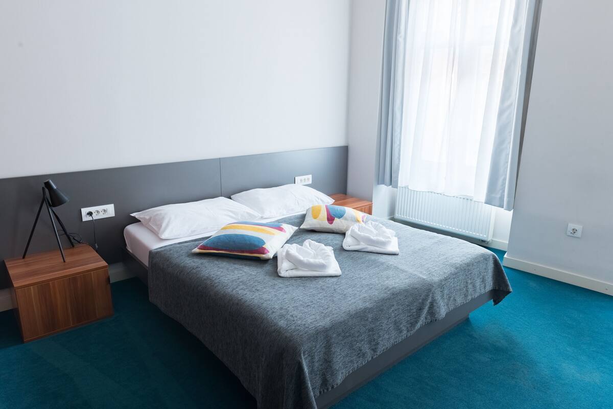 A neatly made bed with a grey bedspread, white pillows, and colorful decorative pillows. There is a wooden nightstand with a lamp beside the bed. The room has a blue carpet and a window with sheer curtains.