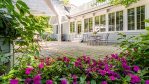 Une terrasse couverte agrémentée d’une pergola blanche, un coin repas avec des chaises et une table en métal, et un jardin où s’épanouissent de magnifiques fleurs roses.