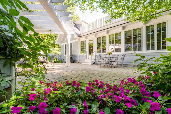 Une terrasse couverte agrémentée d’une pergola blanche, un coin repas avec des chaises et une table en métal, et un jardin où s’épanouissent de magnifiques fleurs roses.