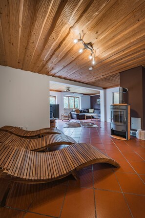 A spacious living area with a wooden ceiling, a built-in wood-burning stove, and a large wooden bench. - Beautiful wooden log house with a first-class view of the mountains! (Bad Goisern am Hallstättersee)