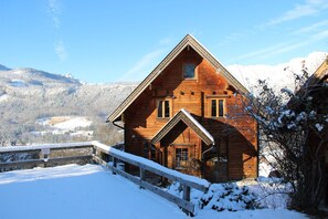 A wooden cabin with a steeply pitched roof, a front porch, and a snow-covered landscape in the background. - Beautiful wooden log house with a first-class view of the mountains! (Bad Goisern am Hallstättersee)