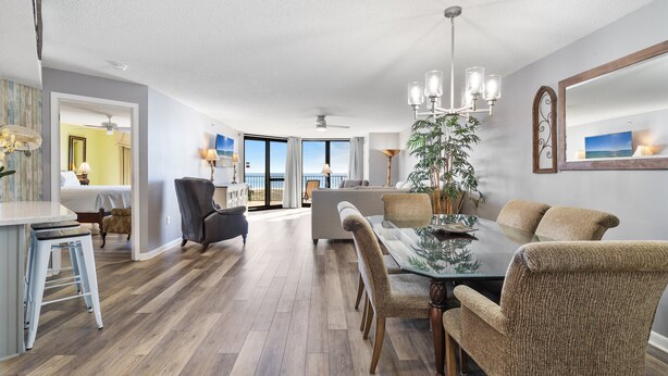 A spacious living area with a dining table, chairs, and a large plant. There is a view of the outdoors through large windows. - Phoenix VI 308 | XL Balcony! | Beachfront Views | Resort Amenities | My Beach Getaways (Orange Beach)