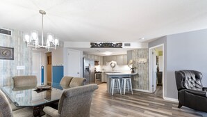 A spacious living room with a glass dining table, comfortable seating, a modern kitchen in the background, and a leather armchair. - Phoenix VI 308 | XL Balcony! | Beachfront Views | Resort Amenities | My Beach Getaways (Orange Beach)
