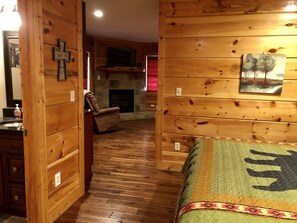 Un interior de cabaña acogedor con paredes de madera, una chimenea de piedra, un sillón cómodo y una cama con colcha.