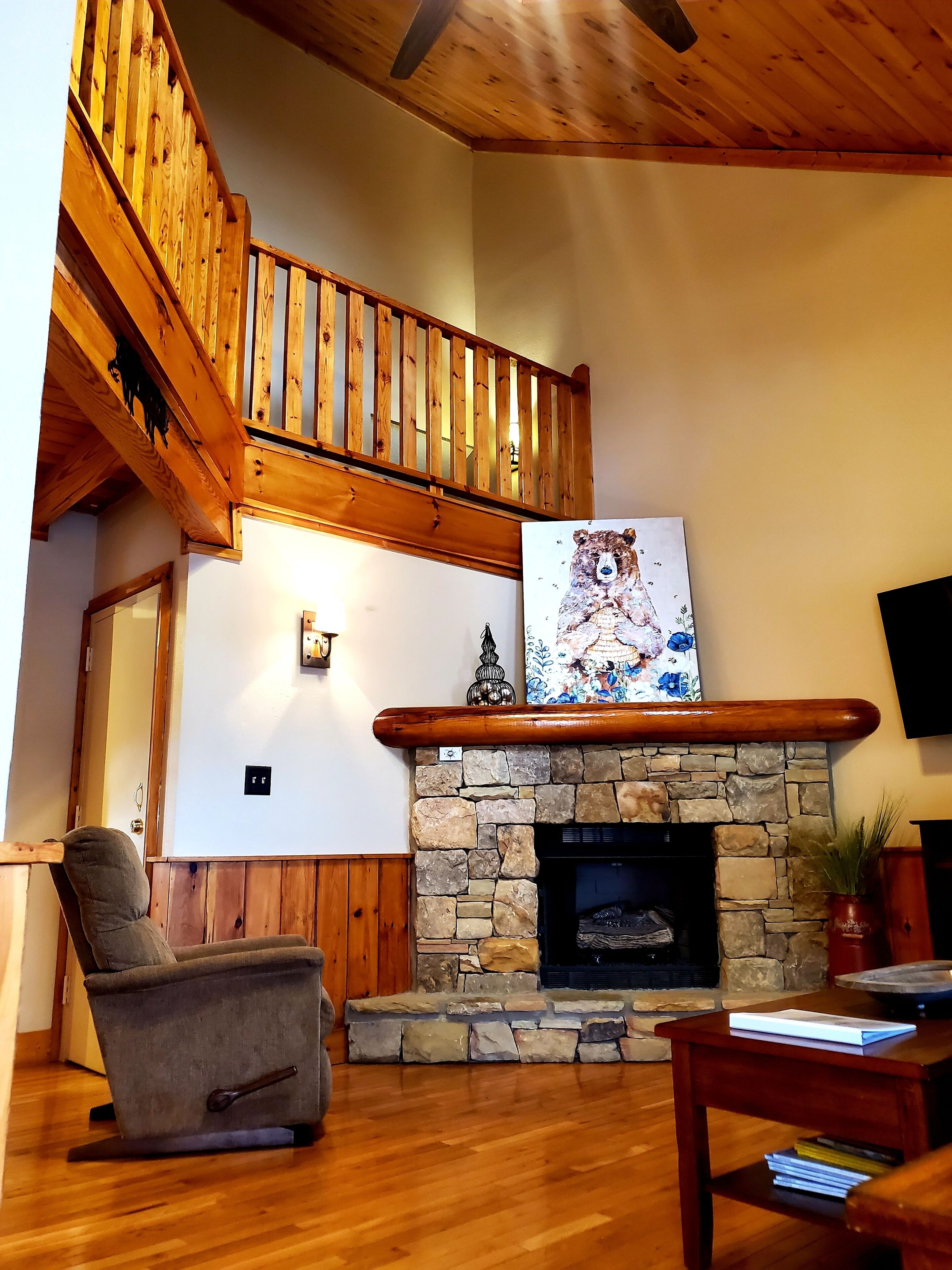 A living room with a stone fireplace, wooden ceiling, and a wooden staircase.