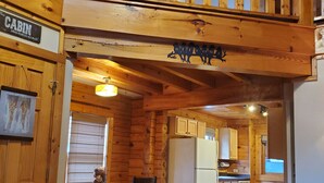 A cabin interior with a dining area, a gray armchair, wooden walls, a kitchen, and a loft bedroom.