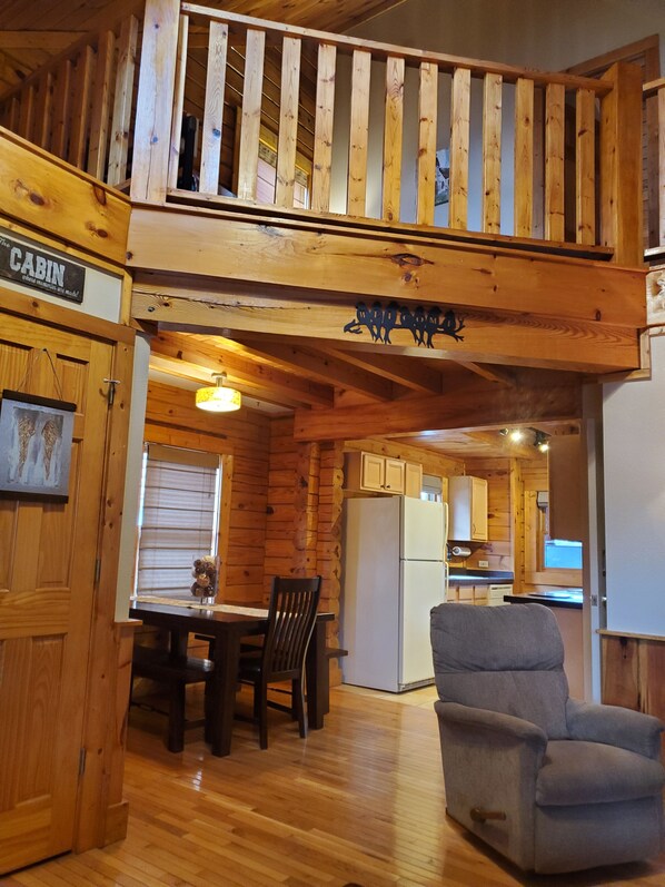 Interior de una cabaña con comedor, un sillón gris, paredes de madera, una cocina y un dormitorio en la buhardilla.