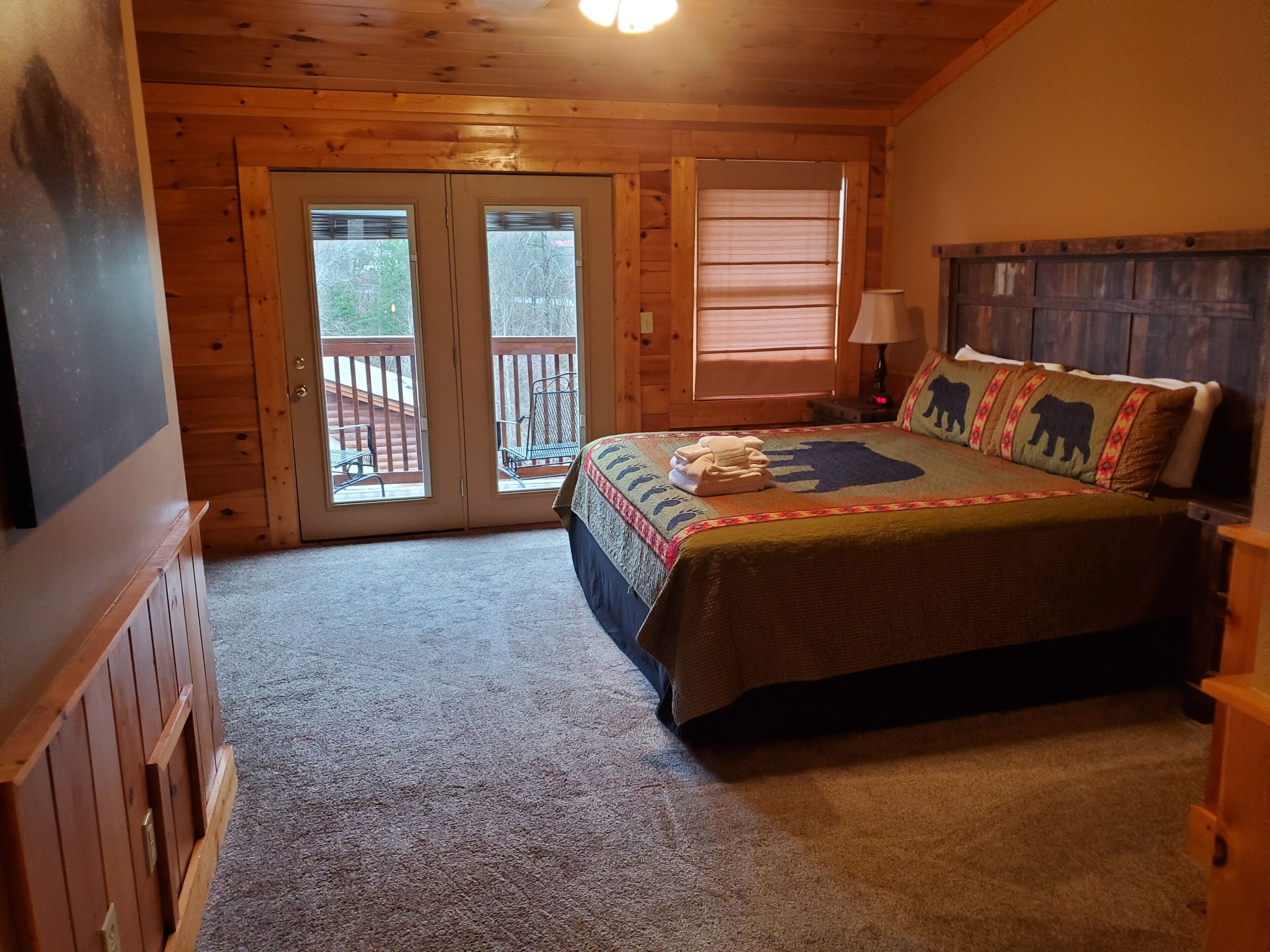 A cozy bedroom with a wooden ceiling, a large bed with a bear-themed bedspread, a nightstand with a lamp, and a sliding glass door leading to a balcony.