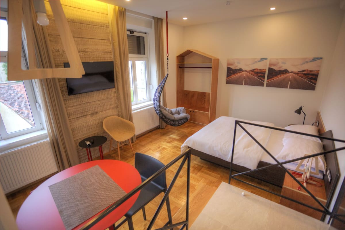 Ein Zimmer mit einem Bett, einem Hängesessel, einem Tisch und einem Fenster mit Vorhängen.