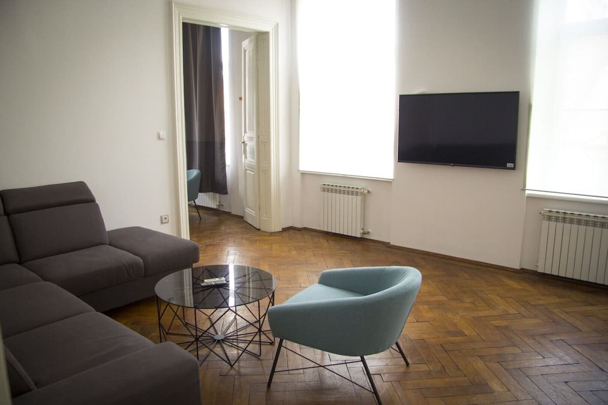 Ein Wohnzimmer mit Sofa, Sessel und Couchtisch. Eine Tür führt in einen anderen Raum, ein Heizkörper ist vorhanden und ein Fernseher ist an der Wand befestigt.