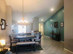 A dining area with a chandelier, a dining table, and chairs. There is a kitchen in the background with a window.