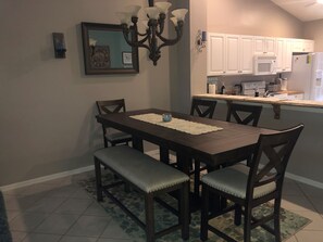 A dining area with a dark wooden table, cushioned chairs, a chandelier, and a kitchen in the background.