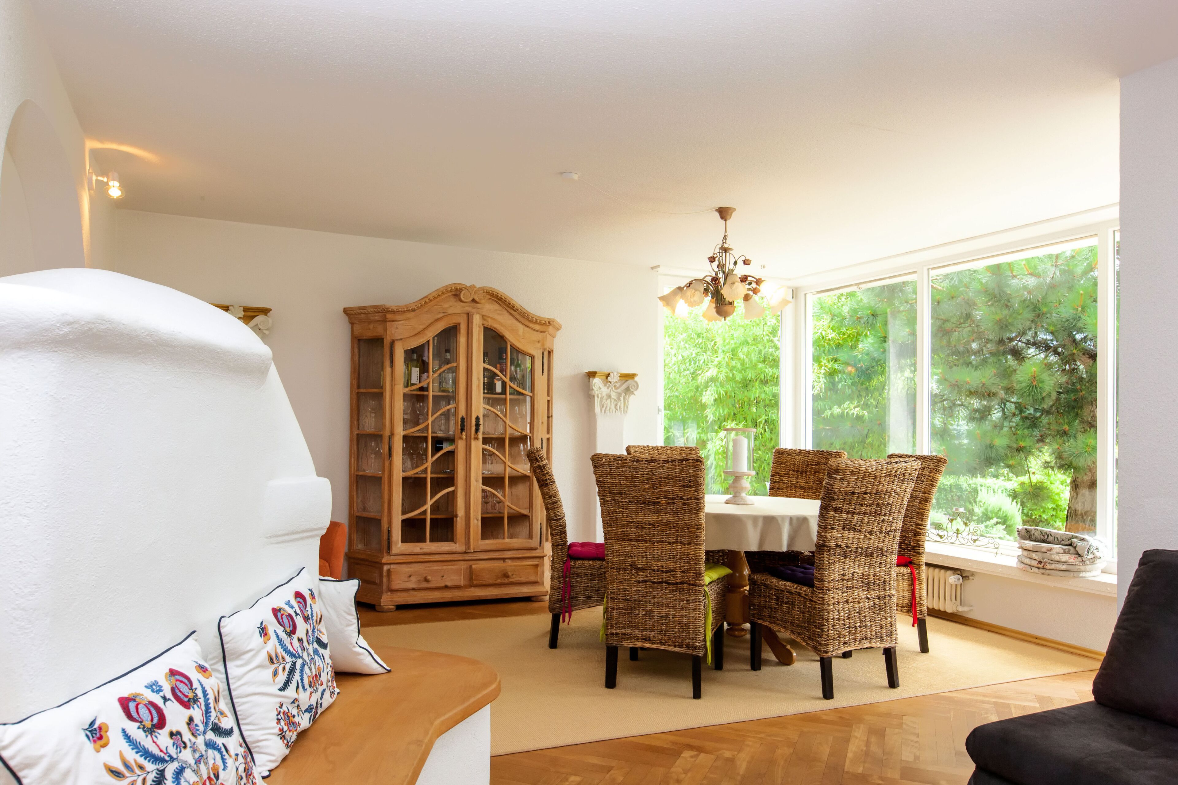 A dining area with wicker chairs, a wooden cabinet, and a view of greenery through large windows.