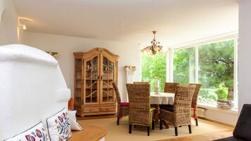 A dining area with wicker chairs, a wooden cabinet, and a view of greenery through large windows.