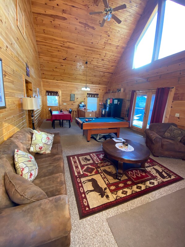 A cozy wooden cabin interior with a pool table, a sofa, a coffee table, and a ceiling fan.