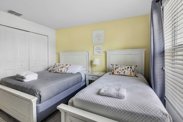 A hotel room with two beds, a white wardrobe, a bedside table with a lamp, and a window with blinds.