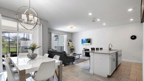 A modern kitchen with a central island, a dining area with a white table and chairs, a living room with a sofa and a TV, and a view of the outdoors through large windows. - Resort Style, close to Disney and other parks Wlking Distance to Oasis Club (Davenport)