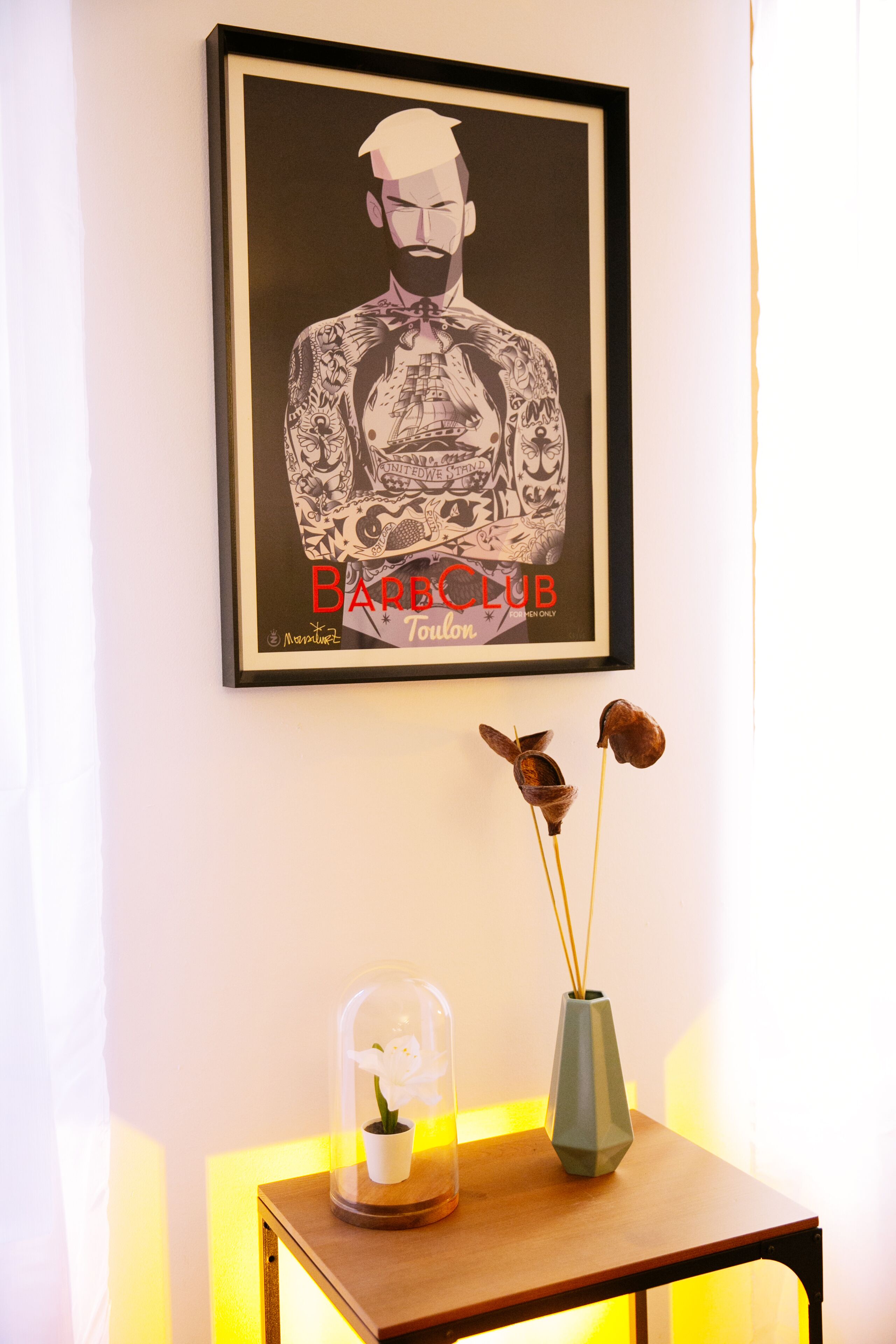 Een ingelijste tatoeagekunstwerk aan een muur, een klein tafeltje met een potplant en een vaas, en een decoratief object dat op een vogel lijkt.