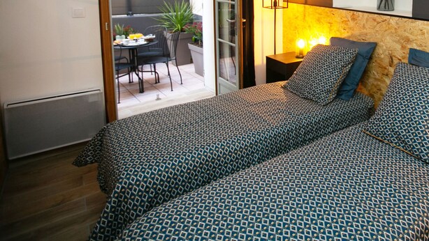 Une chambre à coucher comprenant un lit, une table de chevet, une lampe, un miroir et donnant sur une terrasse extérieure avec une table et des chaises.