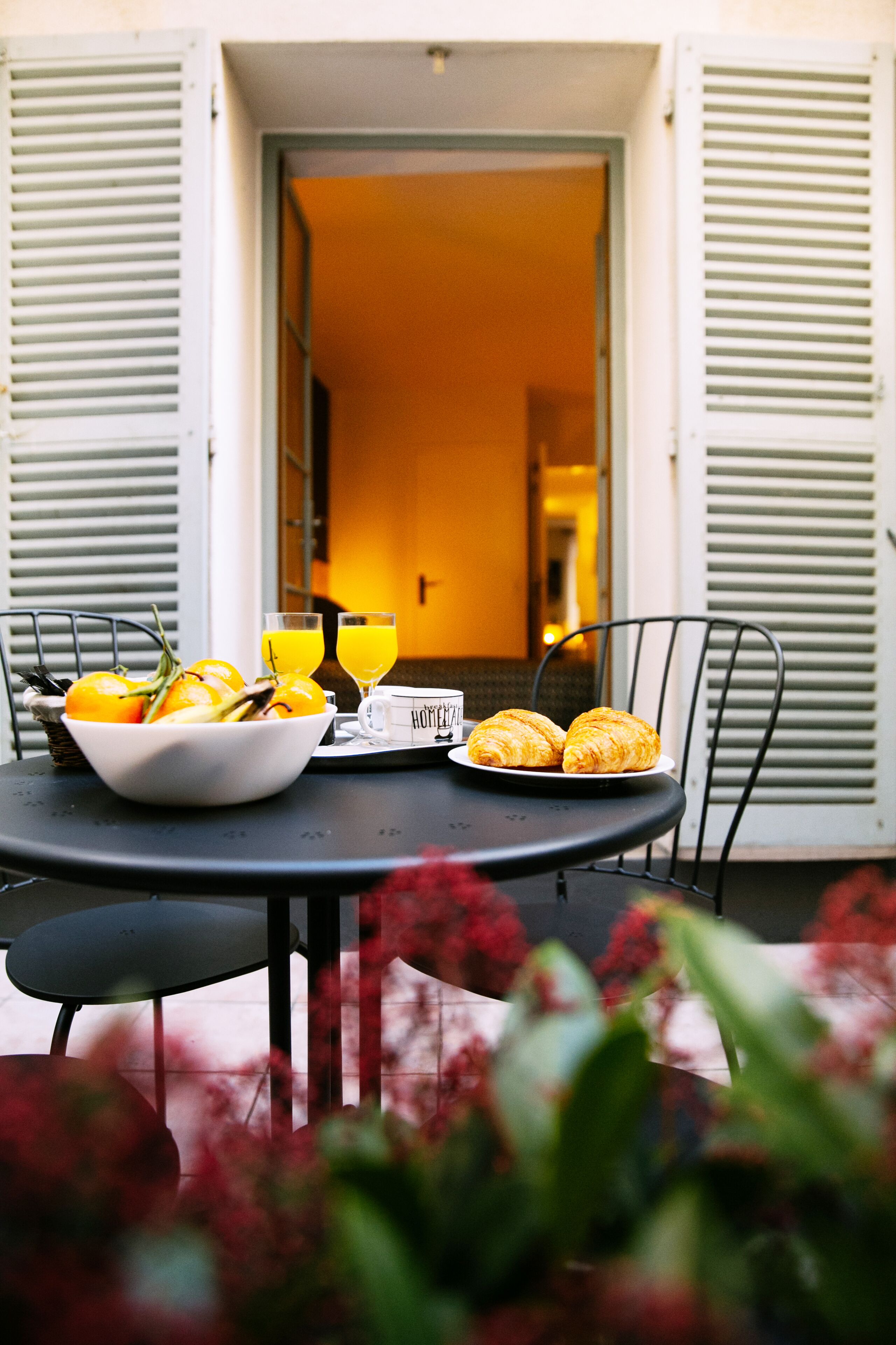 Un tavolo con una ciotola di frutta, due bicchieri di succo d'arancia e un piatto con dei croissant, su un patio esterno con vista su un edificio sullo sfondo.