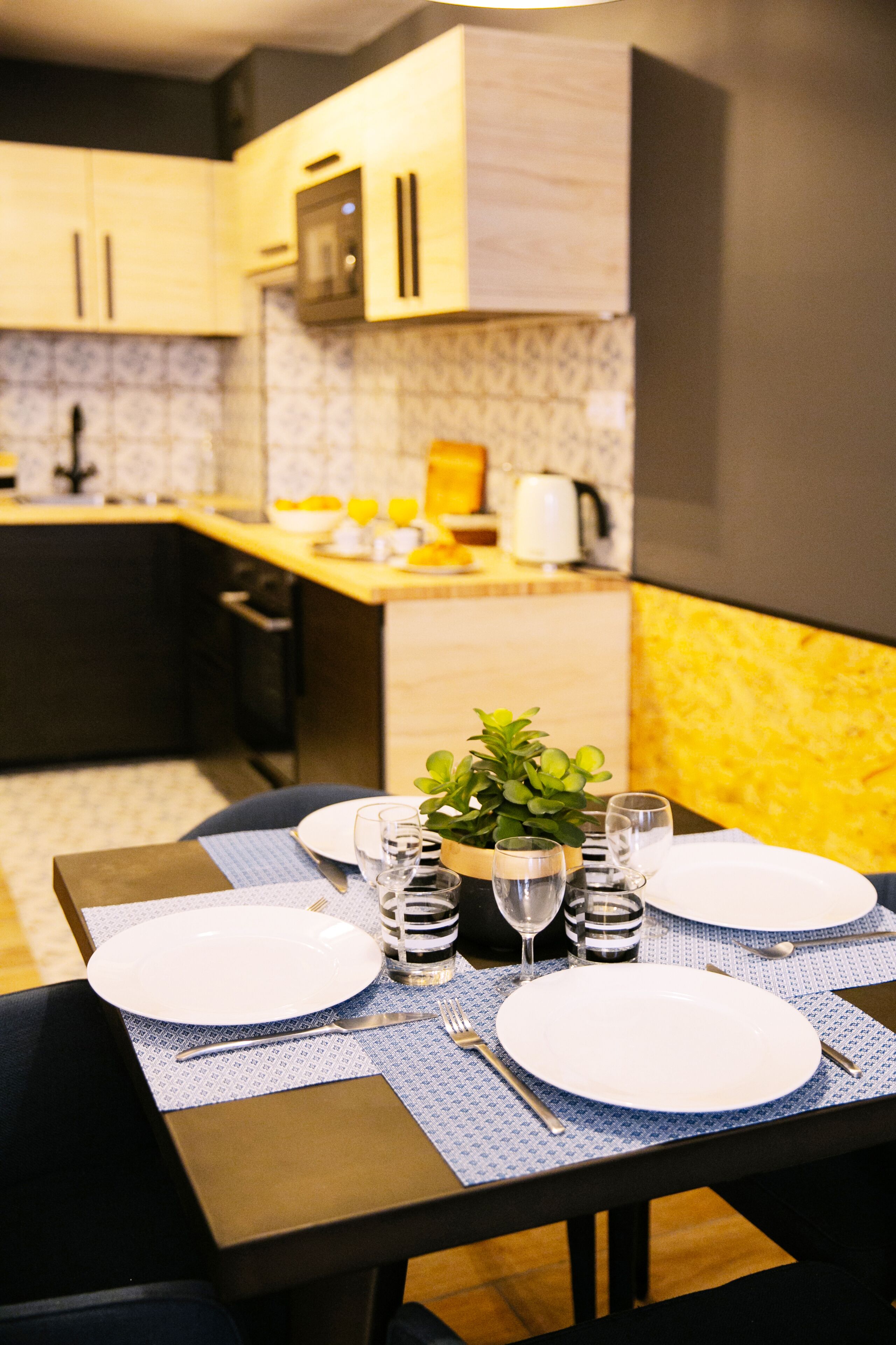 A modern kitchen with a dining table set for two, featuring a blue placemat, white plates, and a small potted plant.