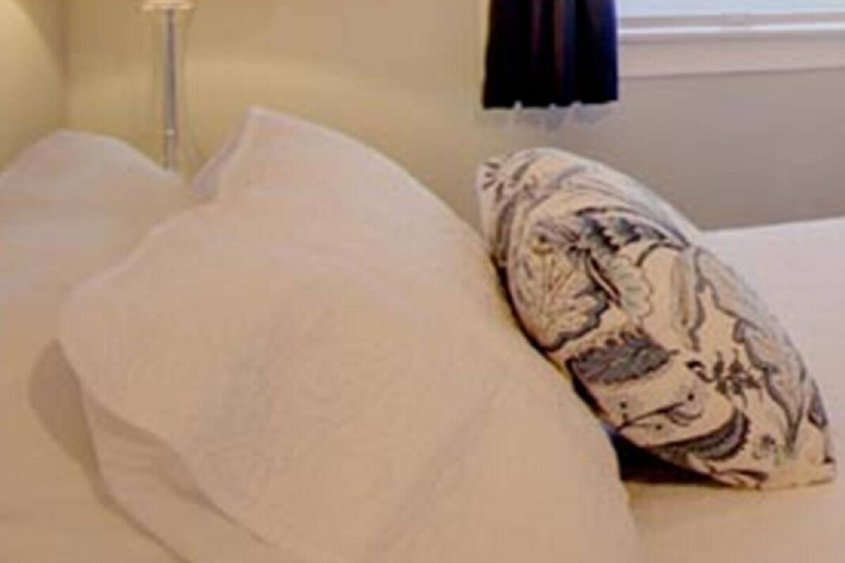 Un lit avec des oreillers blancs et à motifs, une lampe de chevet et une fenêtre avec des stores.