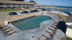 Zona de piscina con sillas de descanso y una pared de piedra, con vista a una playa y al océano.