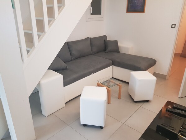 A modern living room with a grey sectional sofa, two white ottomans, a glass coffee table, and a staircase.