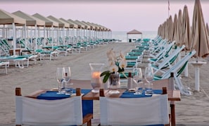 Ein Essbereich am Strand mit Tischen und Stühlen, einer Kerzenarragement und Blick aufs Meer.
