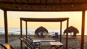 Zona relax in spiaggia con lettini e tavoli sotto un pergolato in legno, con vista sull'oceano.