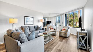 A modern living room with a grey sofa, patterned armchairs, a coffee table, and a TV on a stand. Large windows offer a view of palm trees.