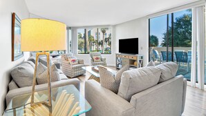 A modern living room with a large TV, comfortable seating, and a balcony with a view of palm trees.