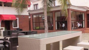 Un área de estar al aire libre con mobiliario de mimbre y una mesa con superficie de vidrio, rodeada por un edificio de ladrillo con grandes ventanales.