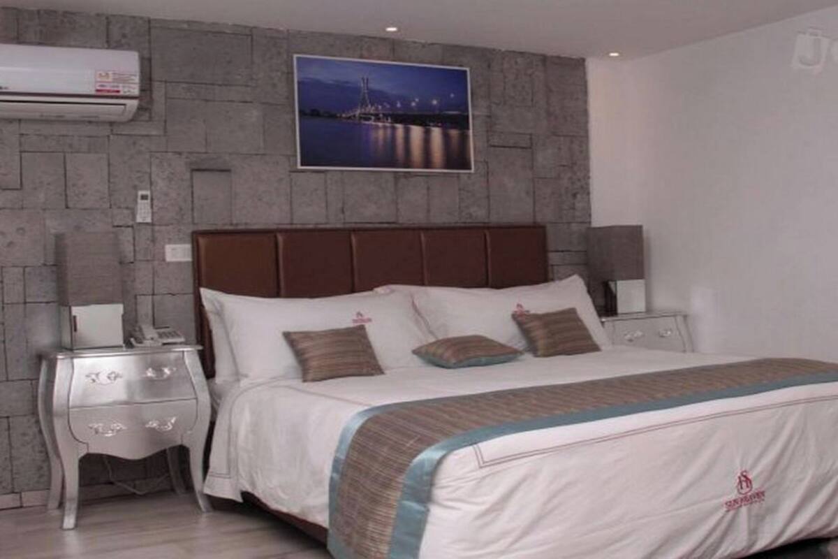 A hotel room with a stone-textured wall, a large bed with a brown headboard, two bedside tables with lamps, and a framed picture of a cityscape.