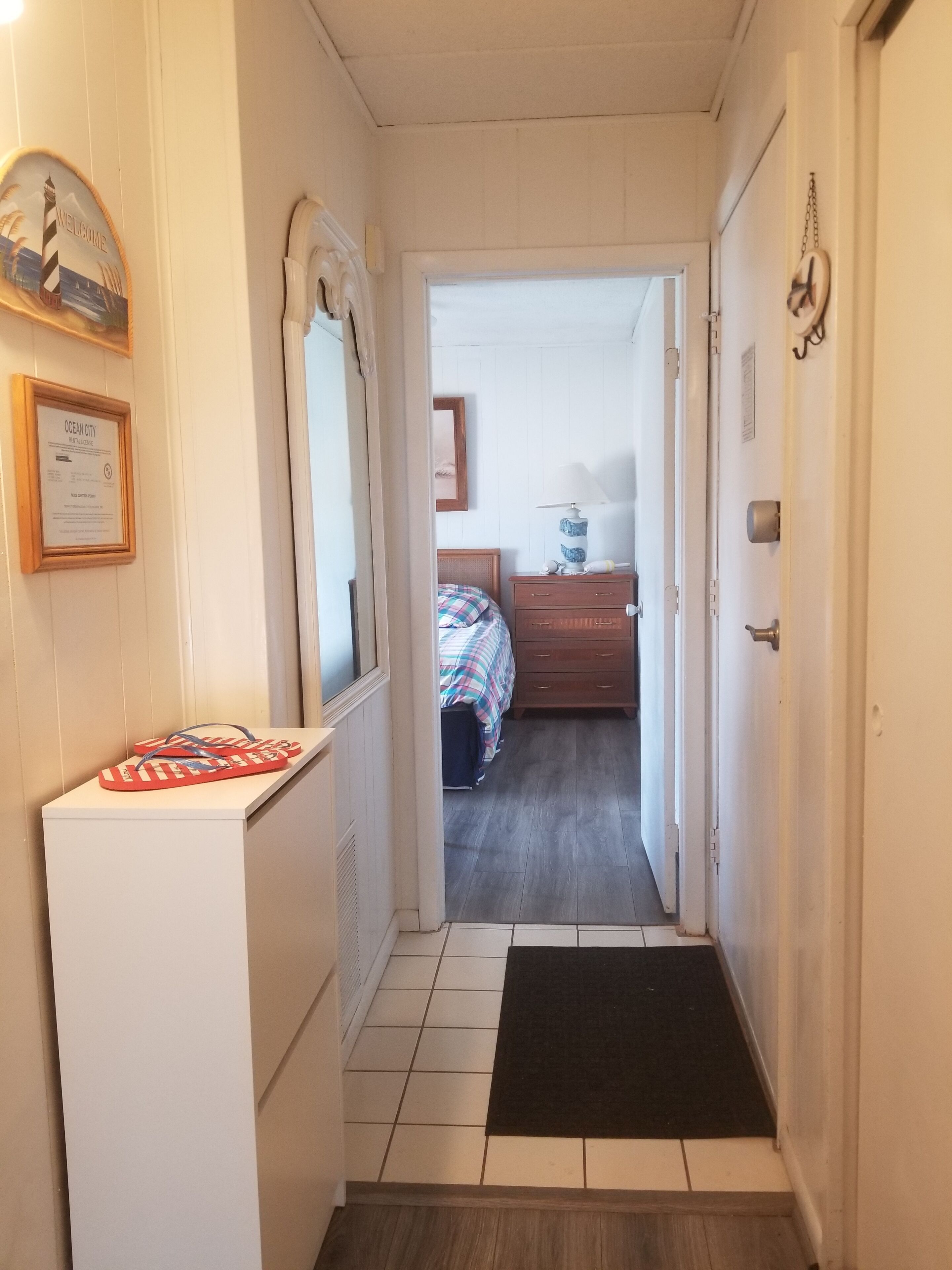 Un pasillo estrecho que conduce a un dormitorio con una cama, un aparador y una lámpara. Se ven un cartel de bienvenida enmarcado y una placa de matrícula en las paredes.
