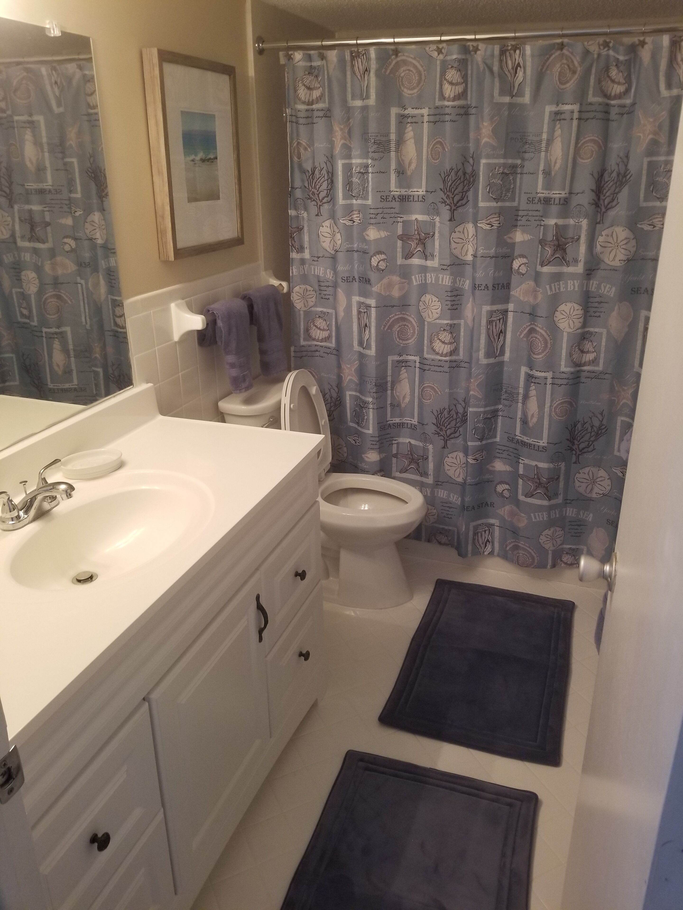 A bathroom with a white sink, vanity, and toilet. There is a blue shower curtain with seashells and sea star patterns. Two dark mats are on the floor.