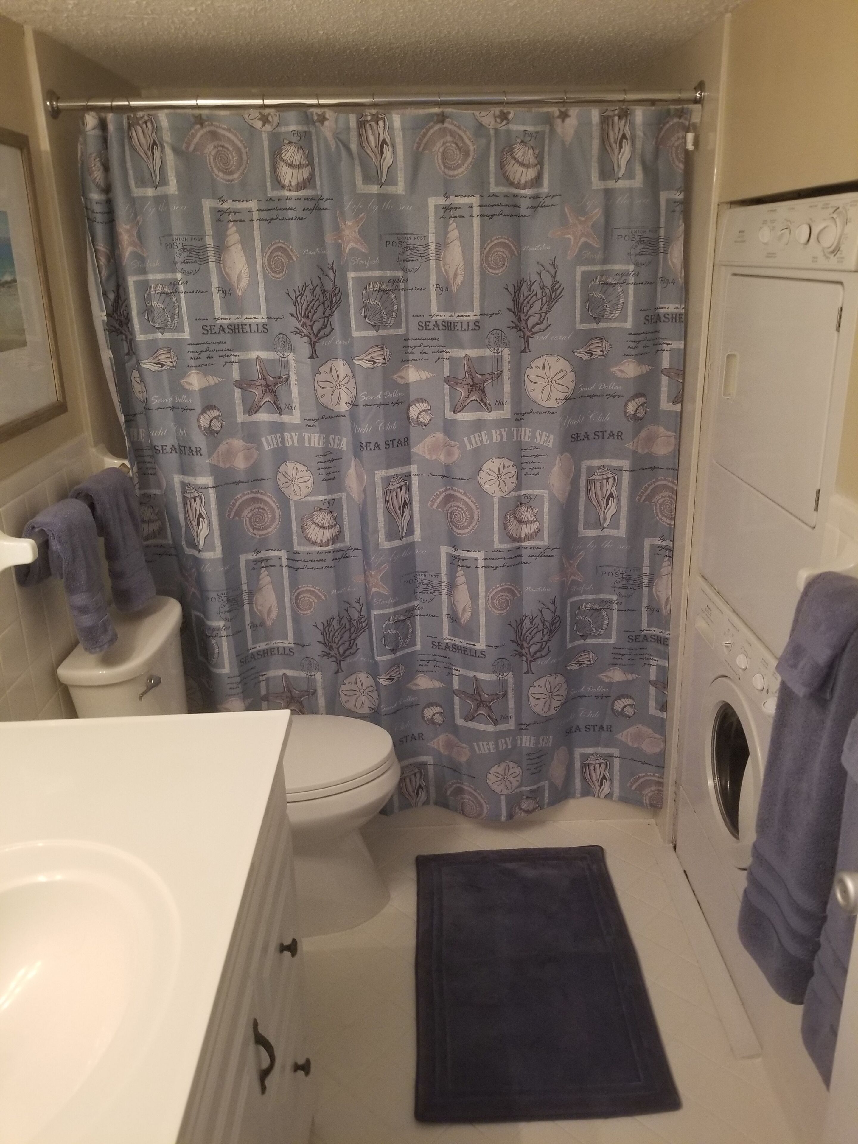 Un baño pequeño con inodoro, lavadora y cortina de ducha con estampado de conchas y estrellas de mar.