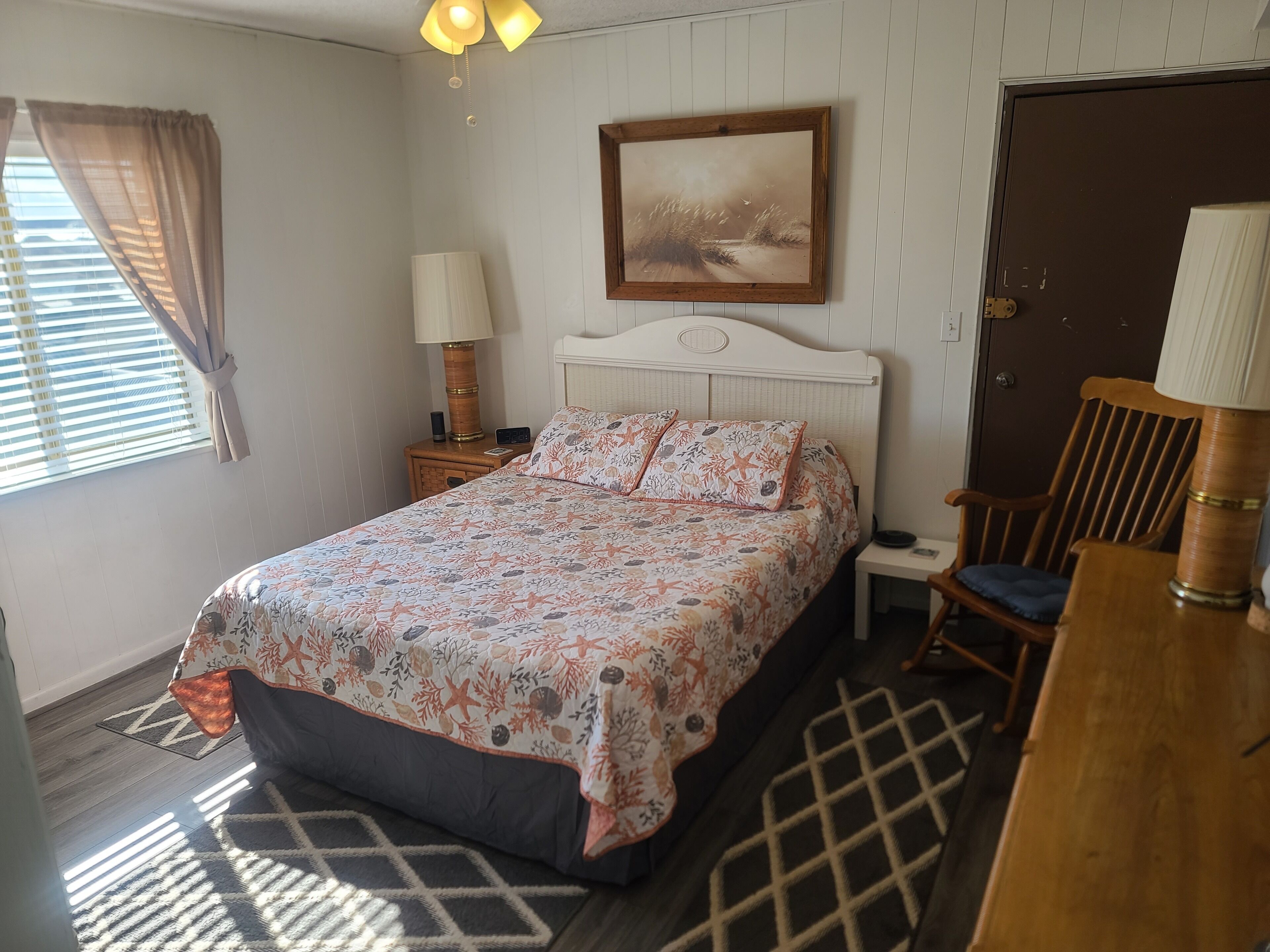 Une chambre à coucher avec un lit, une chaise, une table de chevet, une lampe, un tableau et une porte.