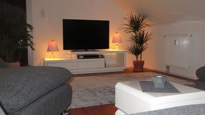 Un salon avec un téléviseur à écran plat posé sur un meuble blanc, deux lampes de chaque côté, une plante en pot et un radiateur.