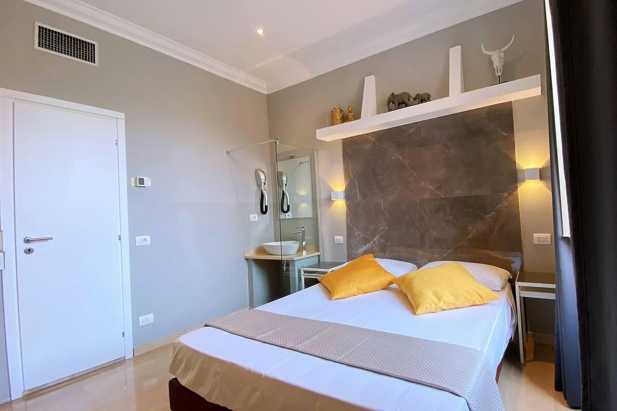 A hotel room with a bed, a bathroom with a mirror, and a shelf with decorative items.