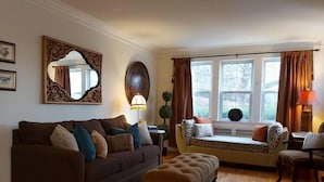 A cozy living room with a brown sofa, an ottoman, a window seat with cushions, and a large mirror on the wall. - 1930's Gingerbread Style - Ambience & Waterviews (Armdale)