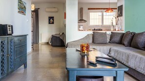Ein Wohnzimmer mit einer Couch, einem Couchtisch und einem Fernseher. Im Hintergrund ist eine Küche mit einem Fenster und einer Pendelleuchte zu sehen.