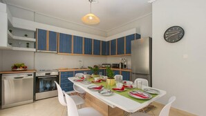 A modern kitchen with blue cabinets, a stainless steel refrigerator, and a dining area set with white chairs and a green tablecloth.