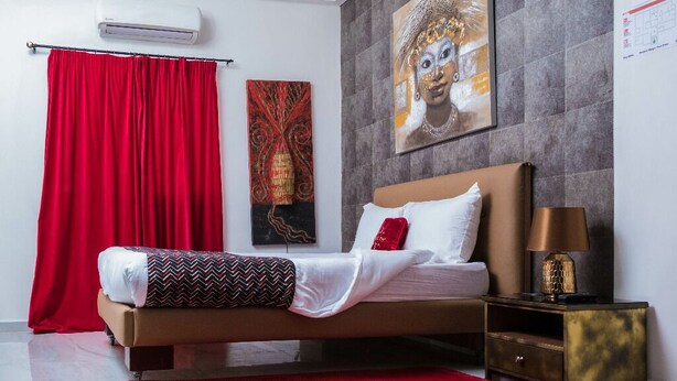 A bedroom with a bed, red curtains, a red rug, a nightstand, and artwork on the wall. - The Key Boutique Hotel (Lomé)