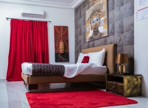 A bedroom with a bed, red curtains, a red rug, a nightstand, and artwork on the wall. - The Key Boutique Hotel (Lomé)