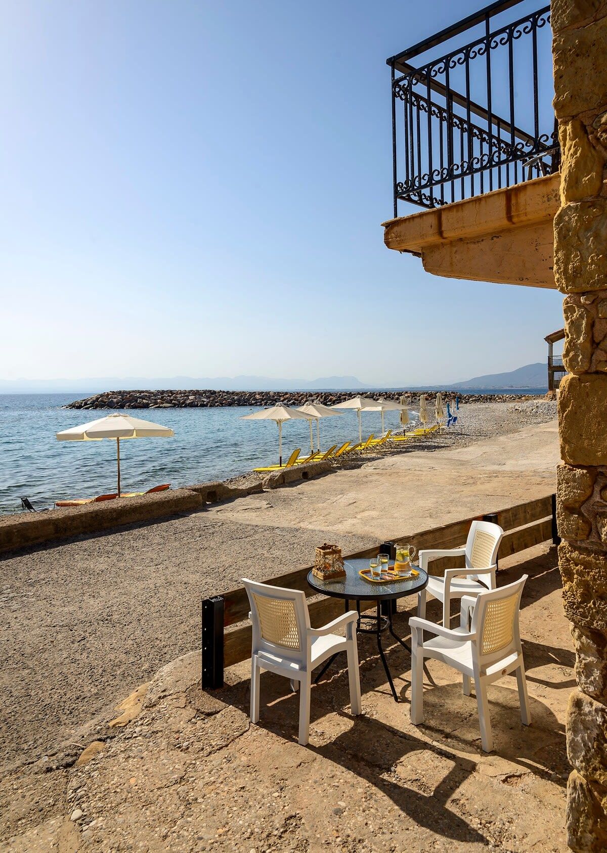 Une terrasse en bord de mer, avec des chaises blanches et une table, d’où l’on a une vue sur la mer et le rivage rocheux.