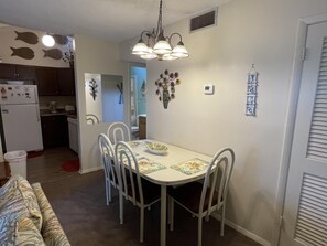 A dining area with a white table, four chairs, and a chandelier. There is a kitchen in the background with a refrigerator and cabinets.