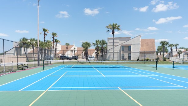 Una cancha de tenis al aire libre con una red, rodeada por una cerca, palmeras y edificios al fondo.